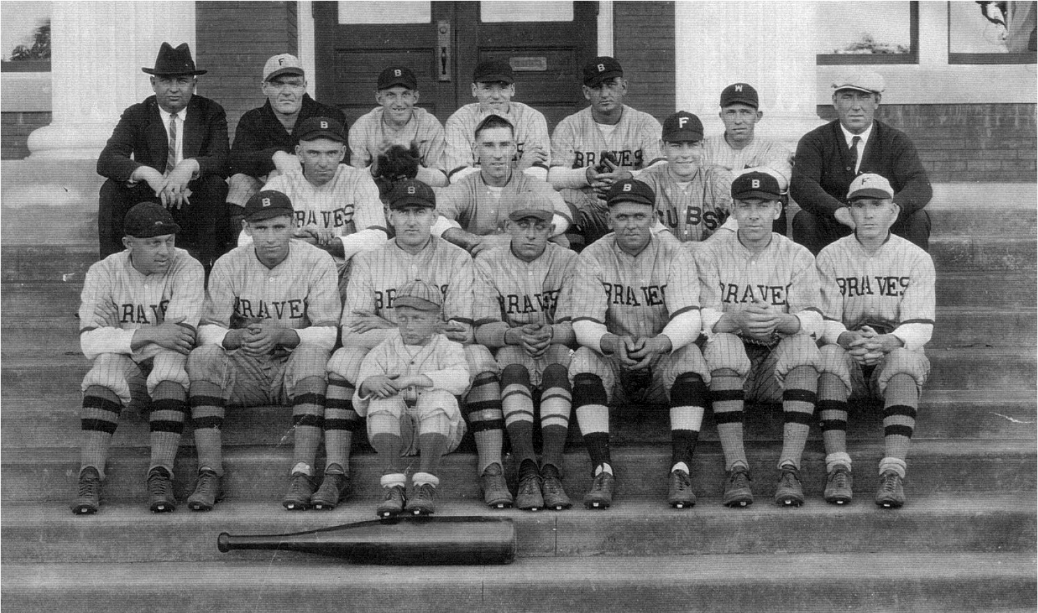 Black-and-white early 20th-century photograph of 17 men and one boy sitting on concrete steps in front of a door to a building, in three rows. Most are wearing light-colored pin striped baseball uniforms with "Braves" on the shirts and tall socks with two wide horizontal stripes. There is a very wide baseball bat on the bottom step.