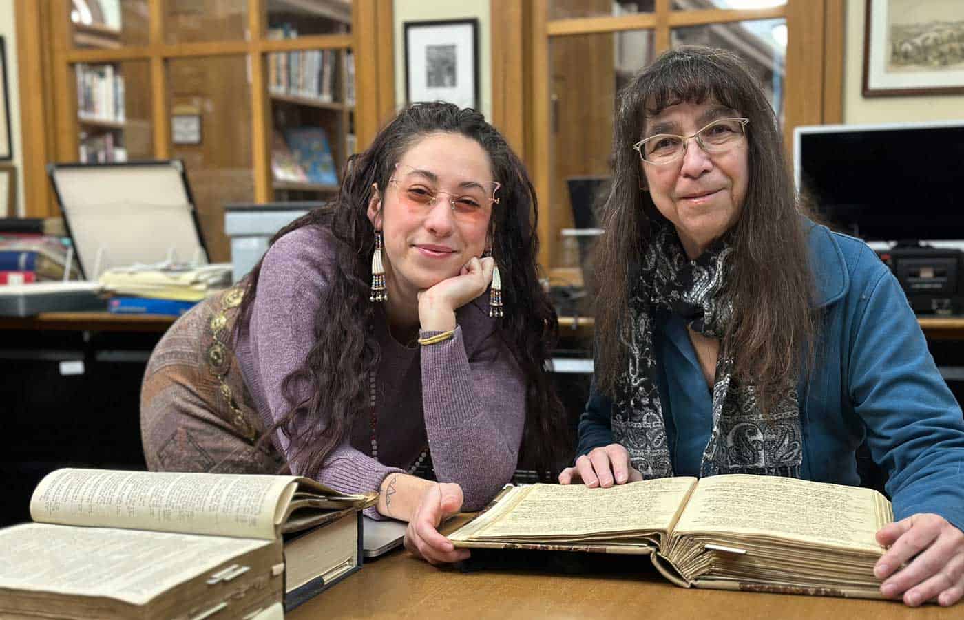 Two women, Dr. Margaret M. Bruchac and Jasmine Rochelle Goodspeed, smile at the camera, with large books on the table before them, in a library setting.