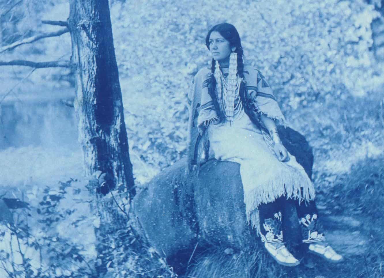 A young woman sits on a large stone beside a pond. She is wearing a fringe-detailed dress, traditional beaded or shell hair ties extending down her braids, and decorated moccasins. She looks off into the distance.
