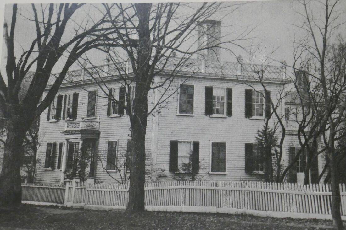 A black and white photograph of a large, two-story historic house with a light-colored exterior and dark window shutters. The home features a flat or low-pitched roof lined with a decorative balustrade and a large, central chimney. A small porch with an ornate upper railing protects the front entrance on the left side of the building. A white wooden picket fence runs along the front of the property, separating the house from the foreground. Several mature, leafless trees stand in the yard, their bare branches partially obscuring the facade of the house.