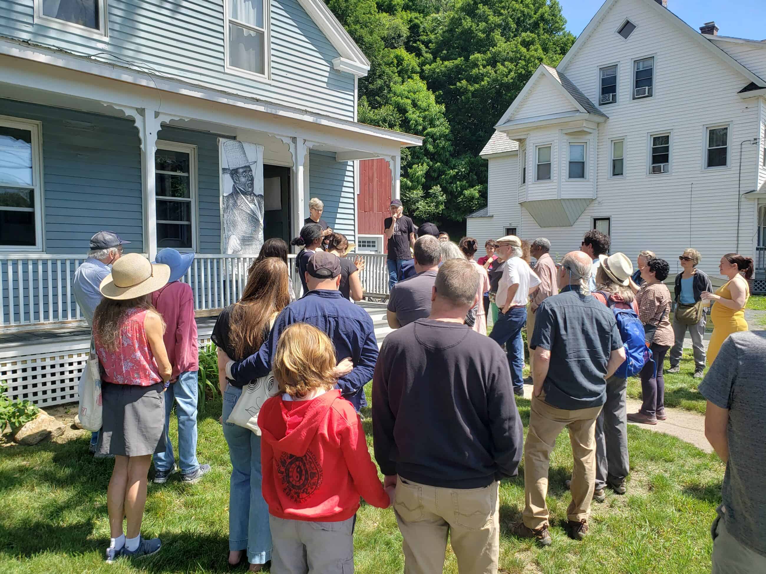 Photograph of people outside of the David Ruggles Center. A banner with a drawing of David Ruggles is hung on the building. A crowd stands looking toward a tour guide or announcer standing in front of the building.