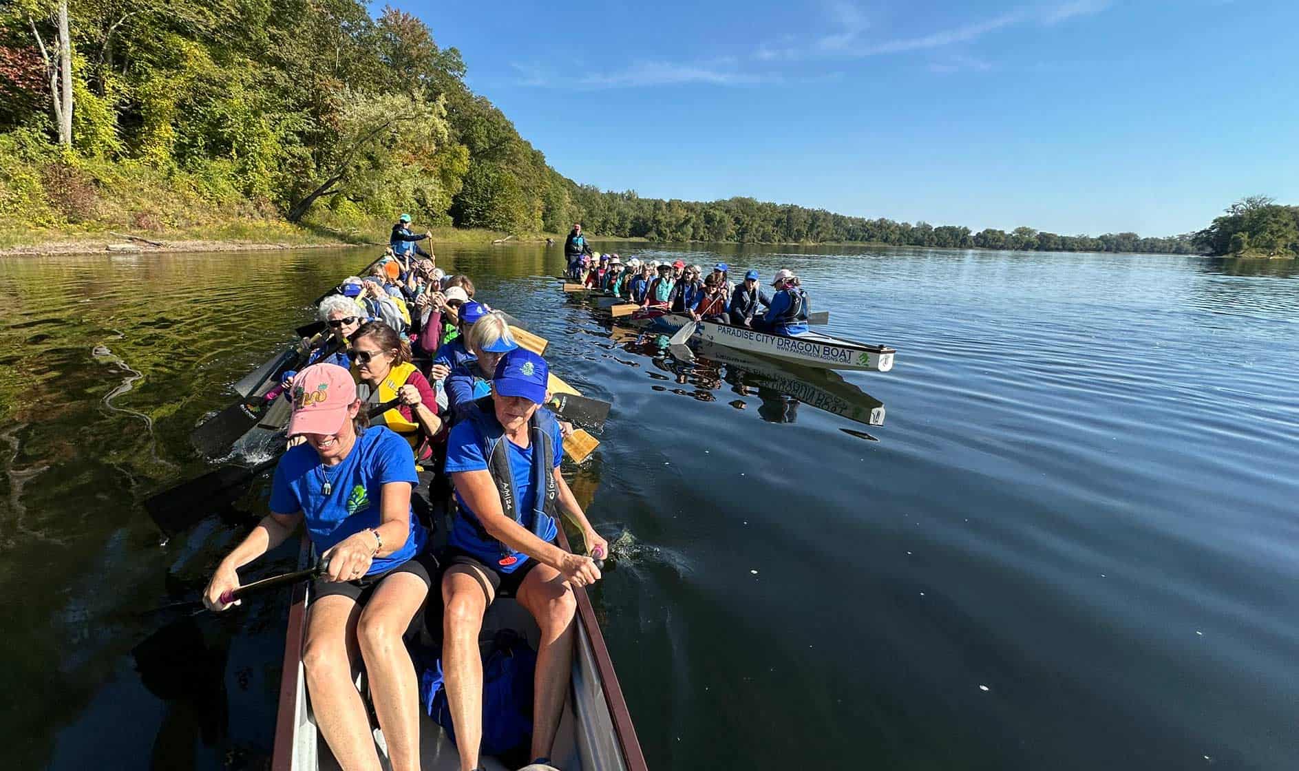 Two long rowboats with about 20 people in each paddling on calm water with green trees on the shore behind.