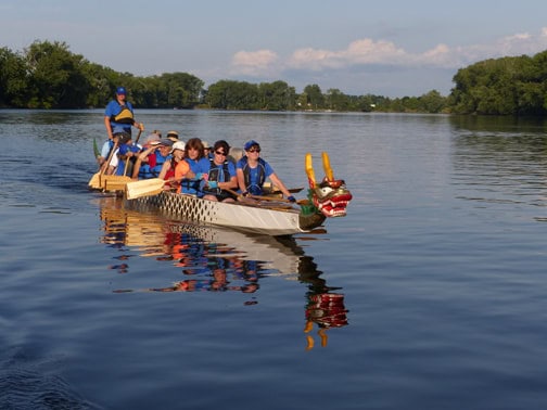 color photo of a long canoe shepaed boat on a river. The boat has a decorative red dragon's head on the front and is full of people, some holding paddles.