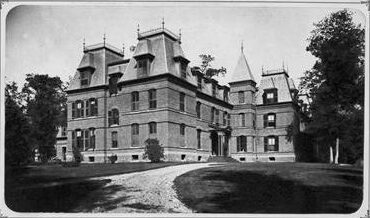 A black and white photograph of a large, multi-story school building with distinctive and ornate cresting along the top edges of the roof. The building features an asymmetrical layout, with several prominent towers or pavilions topped by steep roofs and dormer windows, plus a central recessed entrance with a small porch. A gravel driveway curves across the foreground lawn, leading toward the front entrance.