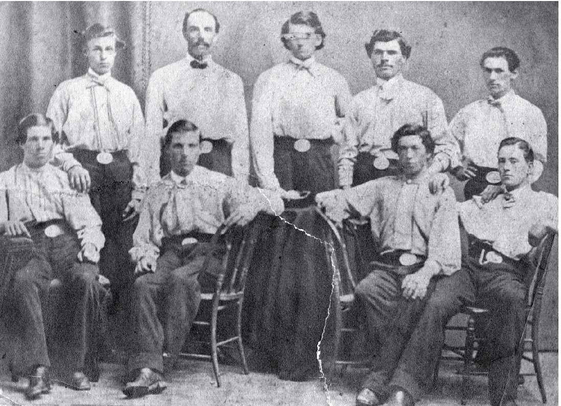 Black-and-white 19th-century photograph of nine men standing and sitting in two rows, looking forward. All are wearing white collared shirts, dark pants, and belts with large oval-shaped silver buckles.
