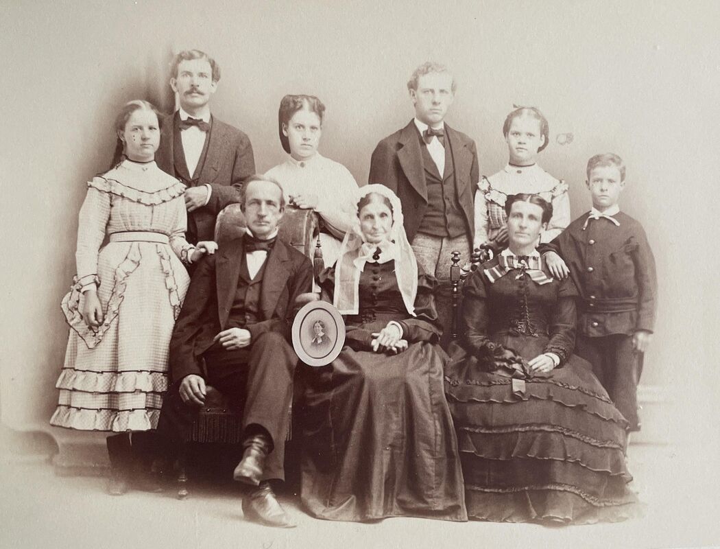 Black-and-white family portrait.
Front row (seated):
Seated Left: A middle-aged man in a dark three-piece suit, white collared shirt, and dark bow tie sits looking forward, holding a small framed portrait in his right hand. Seated Center: An elderly woman dressed in a dark, long-sleeved dress with a white lace collar and a white lace bonnet draped over her head rests her hands in her lap. Seated Right: A woman sits in a dark, tiered gown adorned with intricate details on the bodice and a dark ribbon bow at her neck.
Back row (standing):
Standing Far Left: A young woman stands in a light-colored, long-sleeved dress featuring patterned tiers, layered ruffles on the skirt, and dark boots. Standing Second Left: A man stands tall in a dark suit jacket, matching vest, white shirt, and bow tie, sporting a prominent mustache. Standing Third Left: A young woman stands behind the seated man, dressed in a plain, high-collared light blouse. Standing Third Right: A young man stands in a dark suit jacket, light-colored trousers, a white shirt, and a bow tie. Standing Second Right: A young woman stands in a light-colored patterned blouse-like dress with ruffled detailing around the neckline. Standing Far Right: A young boy stands at the edge in a dark, structured button-up jacket and matching trousers.