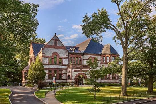 A large, historic Romanesque Revival building made of light stone with contrasting red brick trim around the arched windows and roofline. It features a steep, multi-gabled gray roof, a prominent arched entrance on the ground floor, and a paved walkway leading up to it. The building is surrounded by green lawn, mature trees, and a paved driveway to the left.