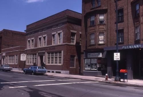 Photograph of the old Northampton Commercial College from 1969. The building is rectangular and made of red-brick, two red brick buildings stand closely next to the building. Two cars can be seen parked on the side of the road.