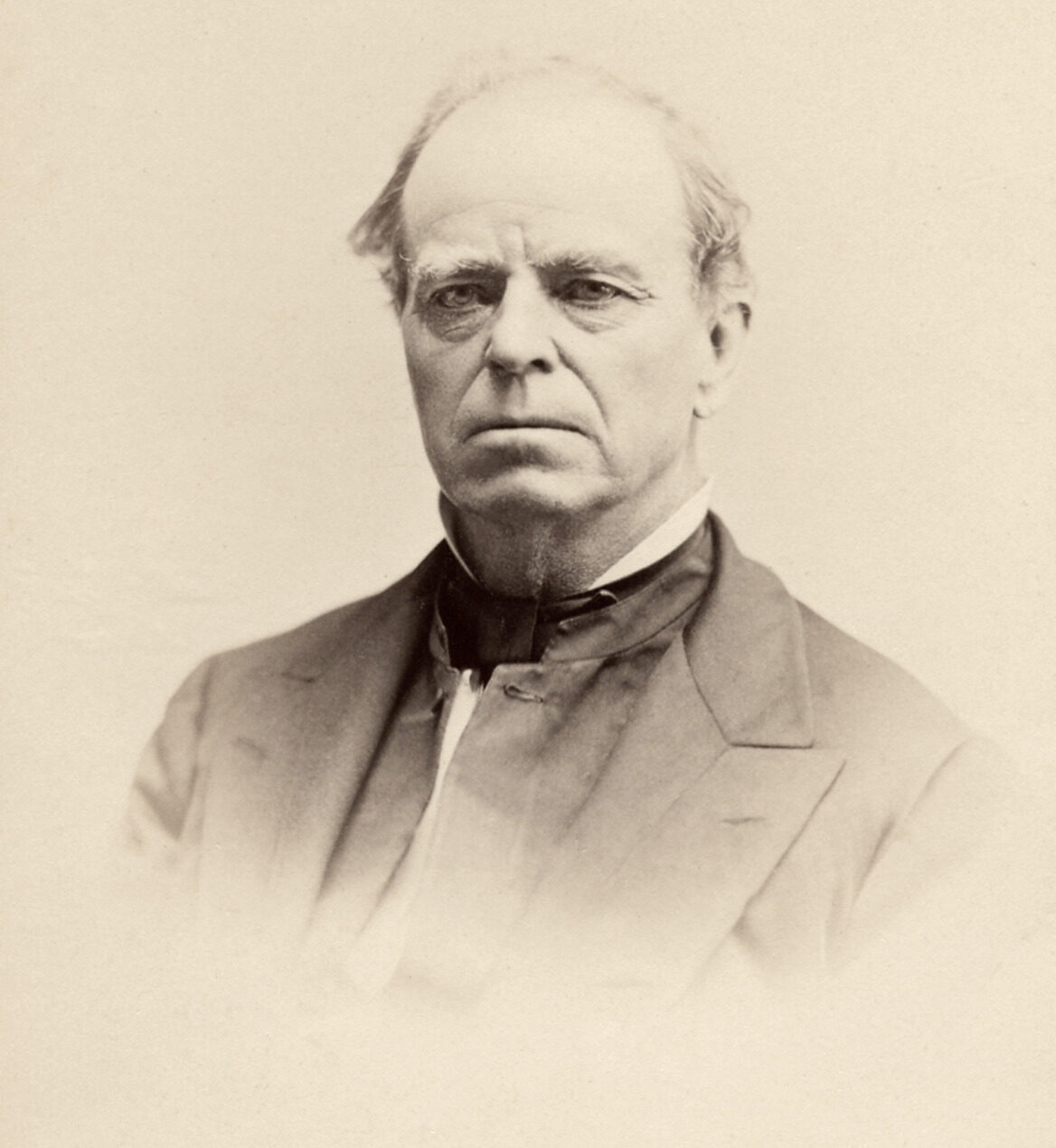 A black-and-white portrait of a man captured from the chest up, He looks directly forward with a serious, solemn expression. He is a mature man with a high, receding hairline, thin graying hair on the sides, and a clean-shaven face with a firm mouth. He wears a dark, wide-lapeled suit jacket over a light vest, a crisp white collar, and a thick, dark cravat tied neatly at his neck. The edges of the vignette portrait softly fade into a clean, light-colored background.