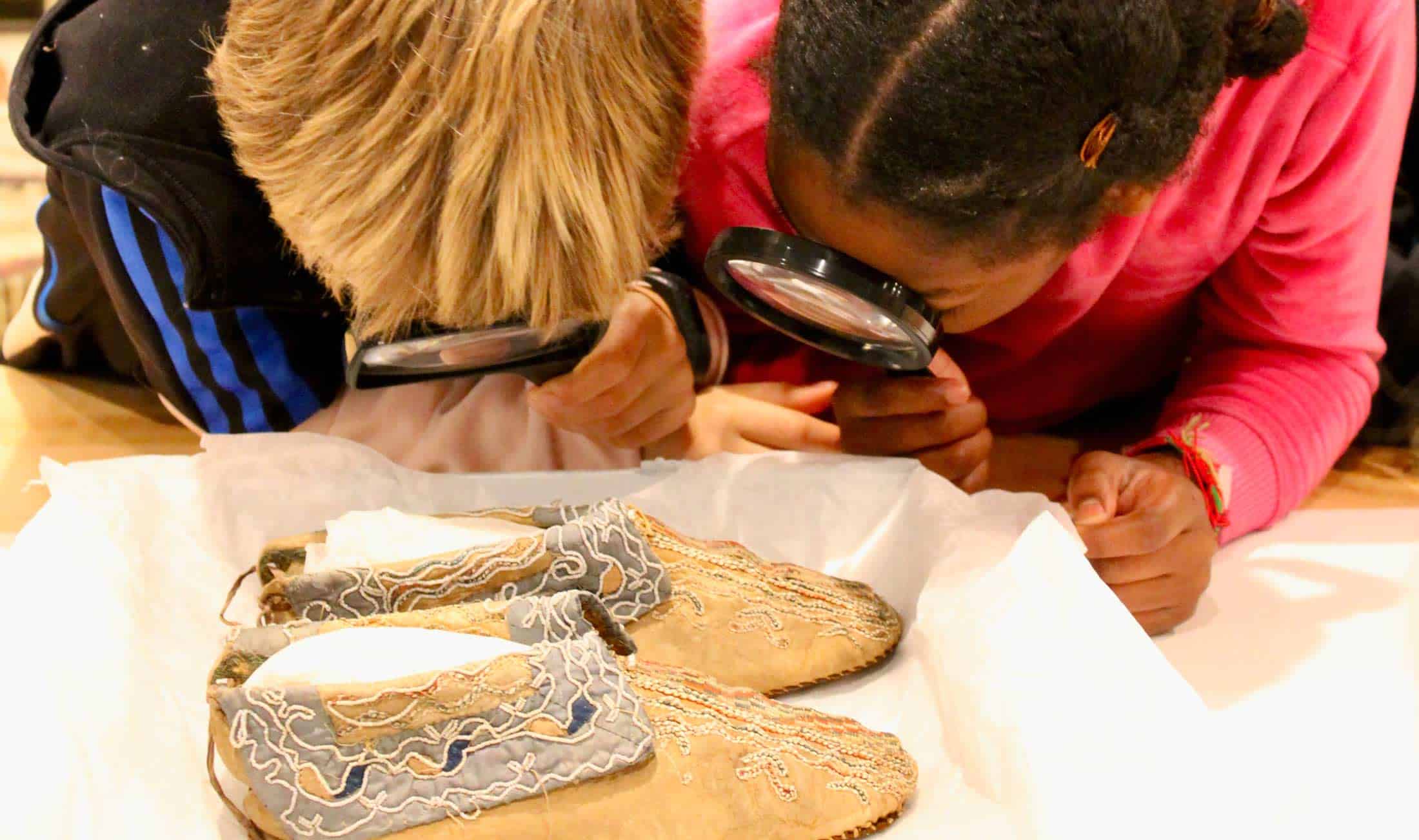 Two children examine a pair of moccasins closely, with magnifying glasses.