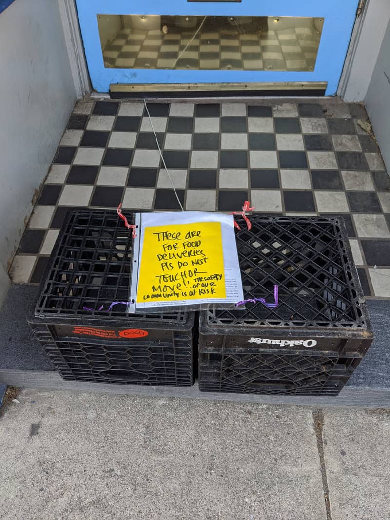 Photo of Store entrance with two milkcrates and sign reading:These are for food deliveries-Pls do not touch or move!  The safery of our community is at risk