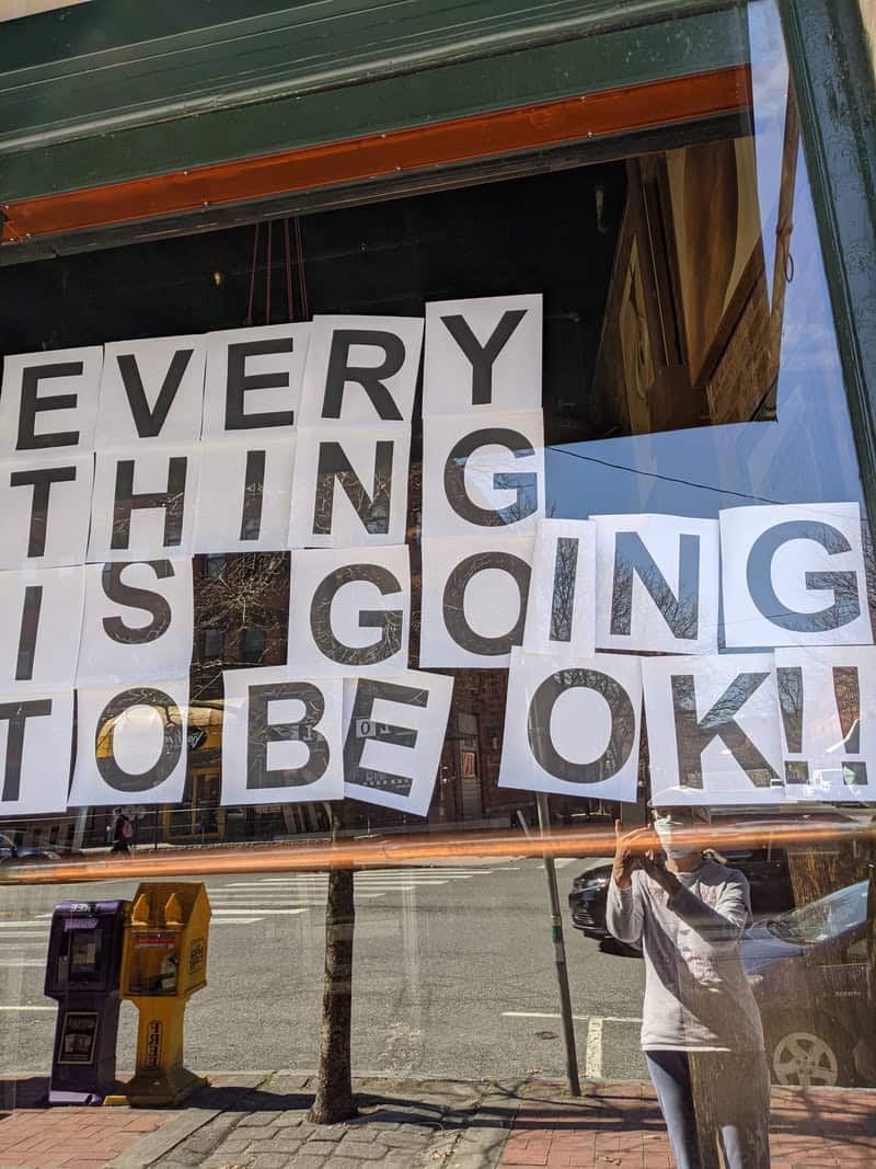 Photo of a store window with sign reading :Every thing is going to be OK!! Reflection of photographer wearing face mask can be seen in window