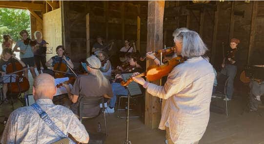 Photograph of about 14 people in a barn playing fiddles, cellos, and other instruments.