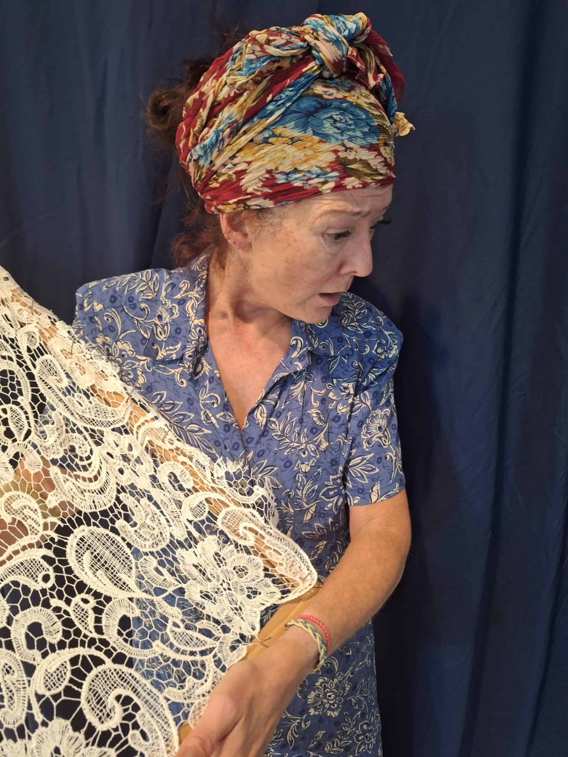 Color photograph of a woman in a blue print dress and scarf wrapped on her head, holding the end of a white lace curtain.