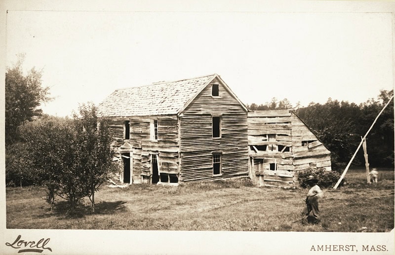 Black-and-white photograph of a wooden house that appears old and rundown. There does not appear to be a door or any glass windows. A man stands infront of the house with his hands in his pockets.