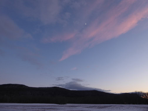 A sunset photo of the sky with a single star and wispy clouds, low hills, and water in the foreground.