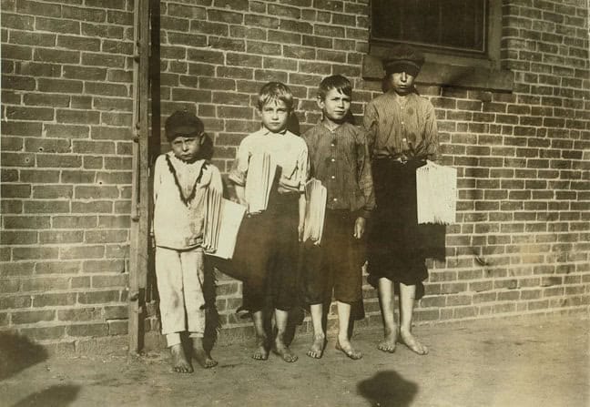 Sepia toned photograph of four young boys standing against a brick wall of a building, in height order, tallest to smallest. Three are holding newspapers under their arms, All are barefoot and wearing worn and dirty clothes. None of them are smiling.