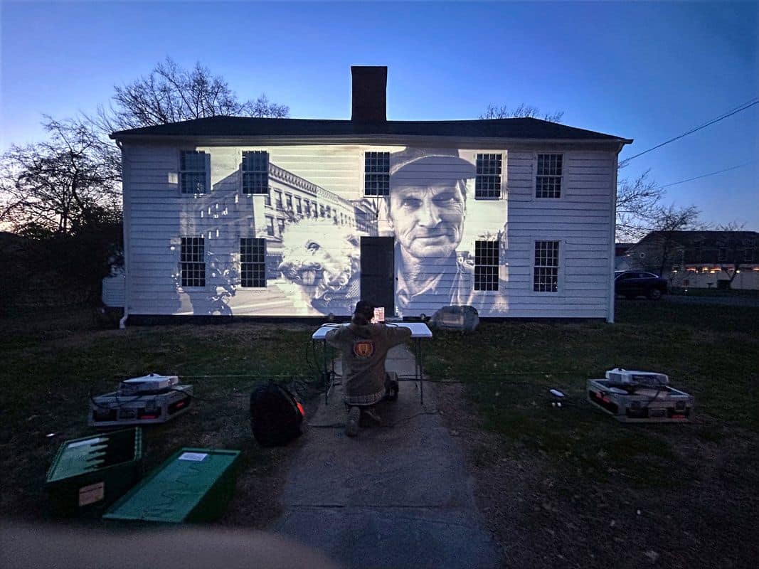 Nighttime photograph of a slideshow being projected onto a white clapboard house. The image is of a man holsing a small dog with a Main Street building in the background
