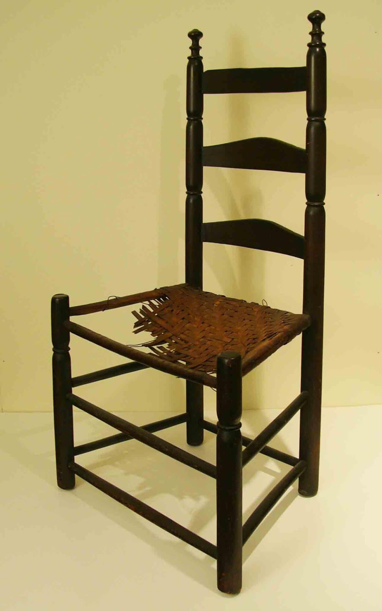 Photograph of a wooden ladder back side chair with a damaged woven ash splint seat.