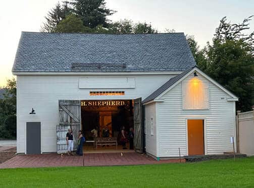 Color photograph of a barn at twighlight with doors open and lit interior shows a large sign inside reading 'H. Shepherd.'