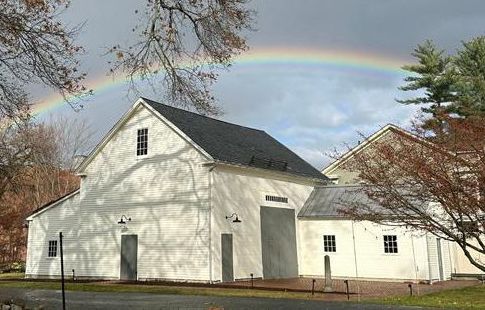 A white, multi-level wooden barn with attached extensions sit under a cloudy sky with a bright rainbow arching over the roofline.