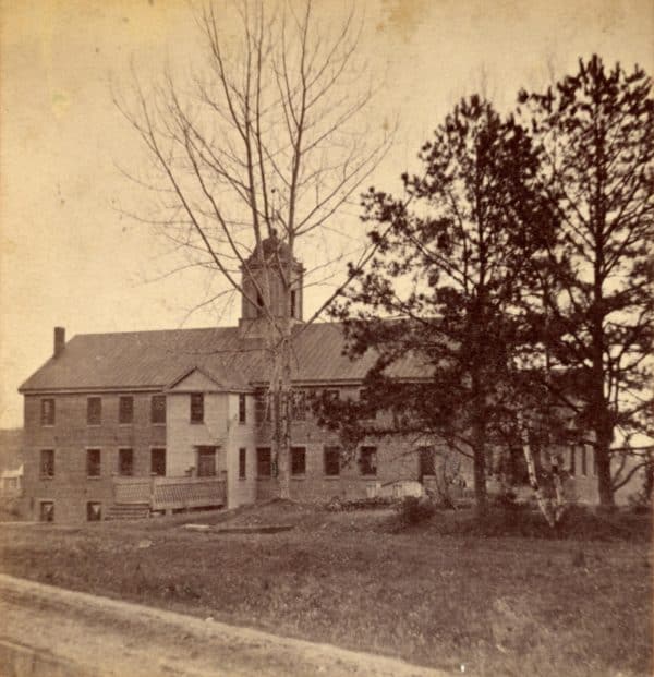 Photograph of the boardinghouse and silk factory of NAEI. The building appears to be brick and is rectangular with a slanted roof and a bell tower. Three trees stand in front of the building.