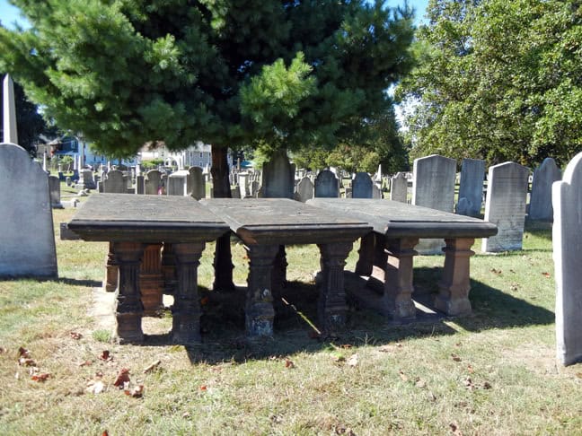 Three stone tables on stone pillars in a cemetery amongst gravestones and trees.