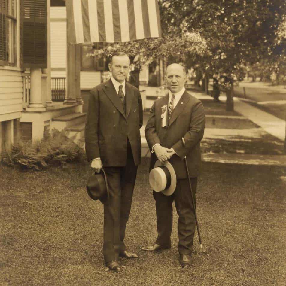 Two men in stand on a lawn in front of an American flag, each with hat in hand.