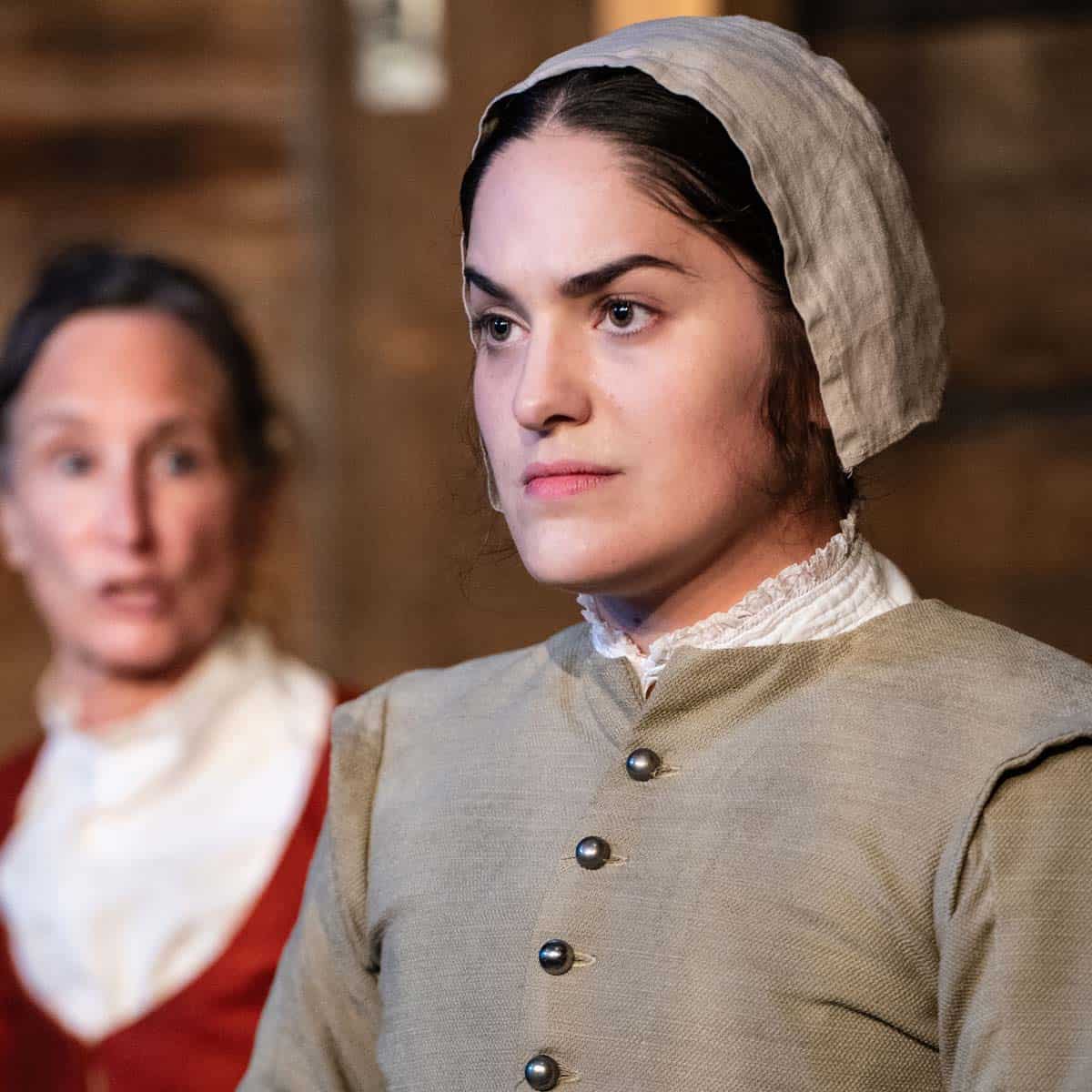 Actors in a play: two women dressed in late-1700s dress, one staring into the distance and the other addressing her in the background.