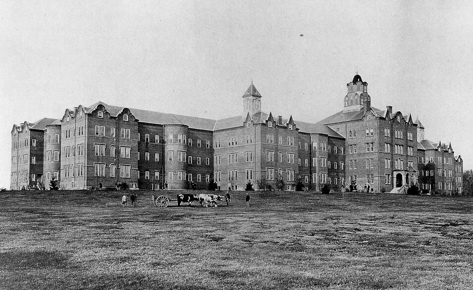 A sprawling and austere brick building on a hill, with cows in the expansive lawn surrounding it.