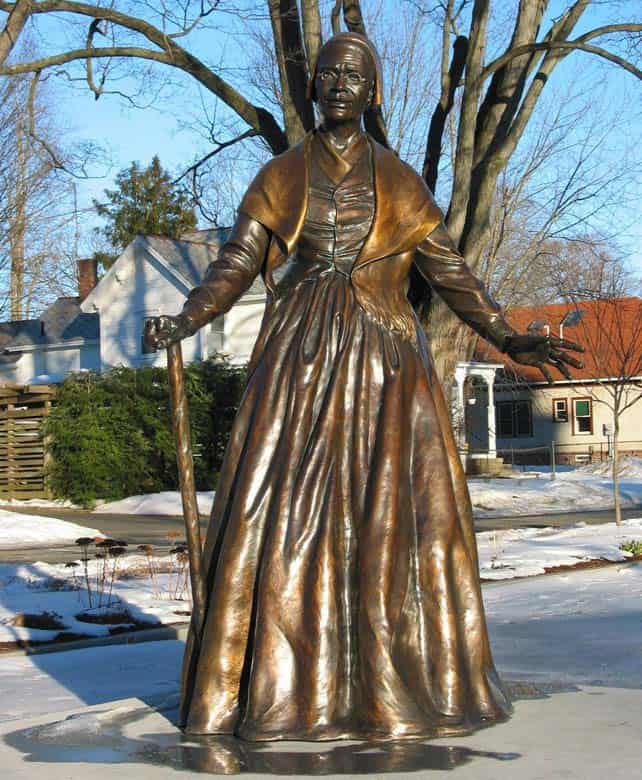 A bronze statue of Sojourner Truth in winter sunshine. She stands with arms slightly outstretched, a cane, and a shawl, in long dress.