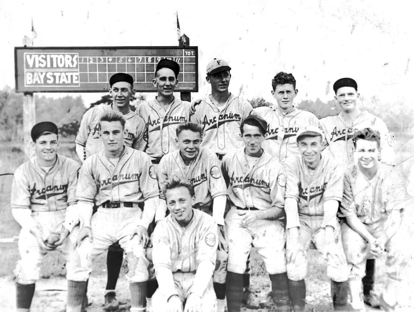 Black-and-white photograph of a baseball team posing in from of a scoreboard. There are 11 men and one teenage boy. Each wears a light-colored baseball uniform with the word "Arcaum" on the front. The scoreboard reads, "Visitors" and "Bay State."