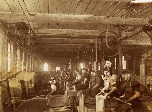 A black-and-white photograph shows a group of male factory workers posing along a busy production line. The workers are arranged in a diagonal line extending from the right foreground deep into the background on the left side of the room. In the immediate right foreground, two young men sit on low wooden stools, one holding a tool or piece of wood across his lap. Behind them, several other men with mustaches and dark work clothes sit or stand at their respective workstations alongside an array of heavy machinery. Large overhead mechanical pulleys, belts, and driveshafts run along the ceiling structure above the workers. To the left, a long row of tall, arched windows lines the wall, letting in a strong, hazy light that creates a bright glare in the background and reflects softly across the wooden floorboards and ceiling beams.