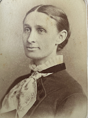 Black-and-white portrait of a young woman in her 20s or 30s, the image cuts off at mid-chest. Her hair is styled in a bun and she wears a ribbon or silk scarf tied around her neck. Her collar is white and puffy, reaching mid-neck, and her dress or coat is black and buttoned.
