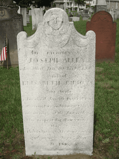 Photograph of a light-colored stone gravestone in a grassy cemetery. The gravestone is typical for the 18th century: Thin and long, with a face or angel design on the top. The top of the marker features a rounded central peak carved with a simple, stylized human face. It is flanked by two smaller, curved shoulders. The face of the stone is weathered with aged, gray textures, displaying worn, engraved text detailing names and dates. In the background, other historic headstones are visible, including a dark gray marker on the left with a small American flag beside it and a reddish-brown marker on the right.