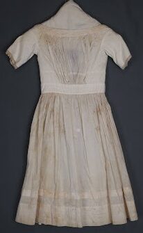 A white dress featuring short sleeves and a wide, gathered neckline. Below the waist, the fabric flows into a full, gathered, knee-to-calf length skirt showing some discoloration and stains consistent with age.