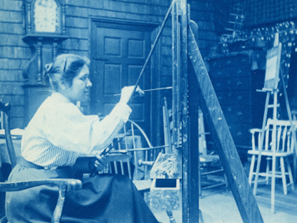 Photograph of a young woman in a white blouse and dark-colored skirt with her hair styled in a bun, sitting on a wooden chair while painting on a canvas. The background appears to be an art studio with chairs and canvas stands.