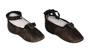 Photograph of dark brown or black weathered baby shoes, each with an ankle strap fastened by a small button. A black fabric ribbon accentuates the vamp, decorated with a small, ornate metallic buckle. The shoes are displayed with white supportive foam inserts inside to retain their structural shape for preservation and photography.