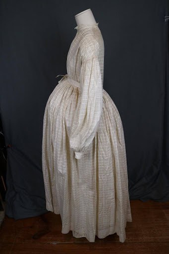 Styled on a mannequin, with a late-term pregnancy bump added. Made of white cotton woven in a checked pattern. The wrapper has a dropped shoulder line and standing collar of white eyelet. The center front opening extends 23" and fastens with cotton covered buttons in the bodice and a double drawstring at the waist. The skirt is cartridge pleated into the wristband more loosely in front and tight in the back. The bottom edge of the bodice back has 2 rows of gathering and there is a self fabric belt attached at the side seams to tie across the front. The sleeves are long and gathered into the dropped armscye and into the cuff at the wrist.