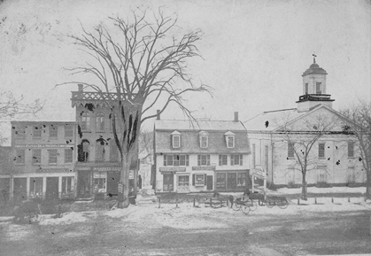 Black-and-white photograph of Main Street. Storefronts and the Edwards Church can be seen. The Edwards Church is white with a slanted roof and a bell tower. Horses and carriages can be seen on the side of the road. There appears to be snow on the ground and the trees in view are bare.