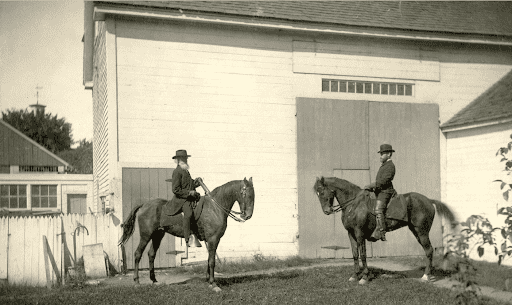 A black-and-white photograph shows two men on horses in front of a white barn. The horseman on the left is older, with a long white beard, wearing a dark suit and hat, and facing right. The horseman on the left is also wearing a dark suit and hat, and facing left. Both horses are dark in color. The barn behind them has a large door. To the left is a white picket fence, and behind it, other buildings and trees can be seen.