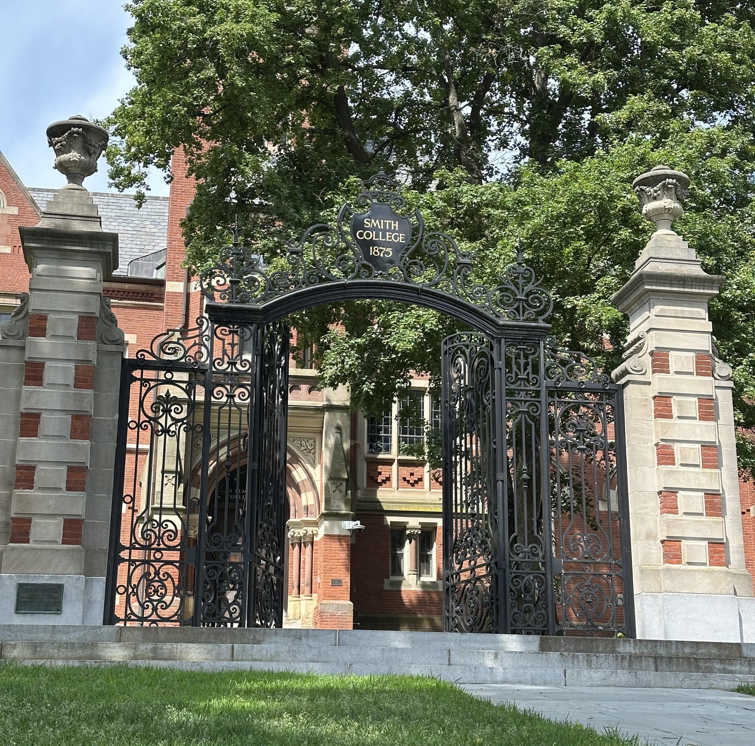 A large, ornate black wrought-iron gate stands wide open at the entrance to Smith College. The top of the gate features a decorative arch with a central black oval sign that reads "SMITH COLLEGE 1875" in goldish-yellow lettering. Flanking the gate are two tall, symmetrical square pillars built from alternating layers of red brick and light gray stone, each topped with a decorative stone urn. Behind the open gateway, a historic brick campus building with arched doorways and multi-paned windows is partially visible, shaded by dense, green trees. The photo is taken from a low angle, showing a stone walkway and a patch of green grass in the foreground under a bright, slightly cloudy sky.