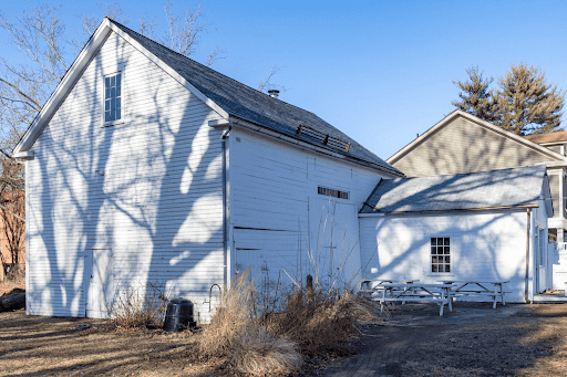 A large white barn with horizontal siding features a steep slanted roof and large sliding barn doors on its side. Prominent bare tree branches cast dark shadows across the left side of the structure. To the right, a smaller, single-story white wing is attached. In front of the barn is a patio area with several white picnic tables. The buildings sit on a grassy yard with patches of dry brush. A portion of another house is visible in the background on the far right.