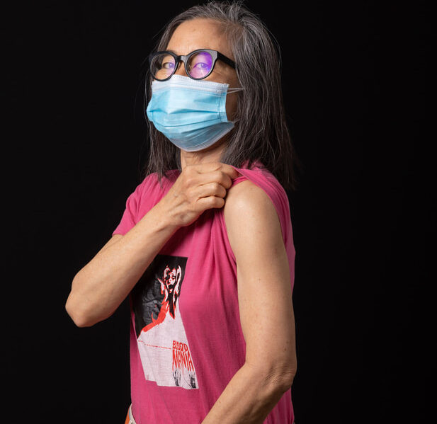 A middle-aged woman in glasses and a surgical mask looks at the camera, showing off her shoulder (the location of her vaccine shot).