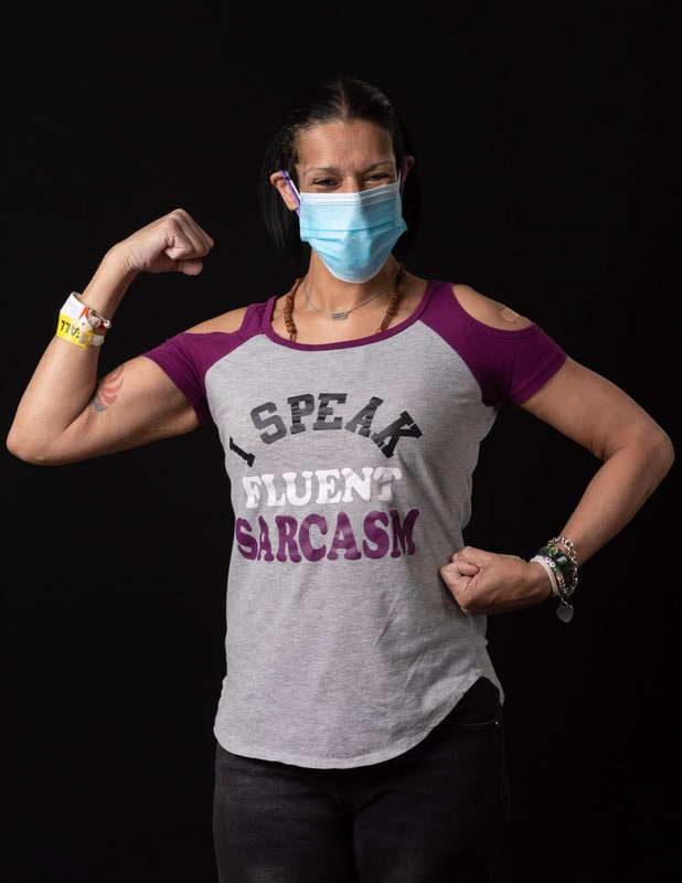 photo of woman wearing face mask and grey and purple shirt reading 'I Speak Fluent Sarcasm' with fist raised